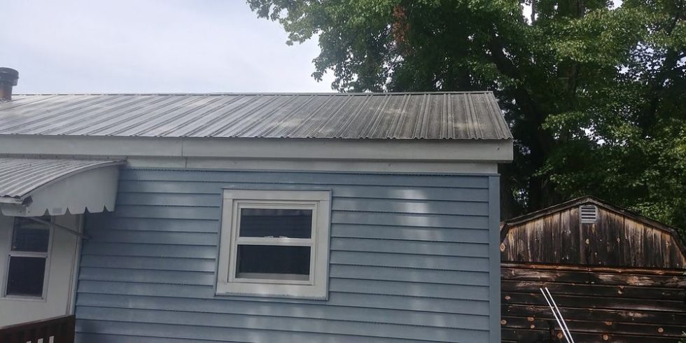 Roof of a blue house showing signs of wear, with a clean, well-maintained appearance after pressure washing, surrounded by trees and a wooden shed in the background.