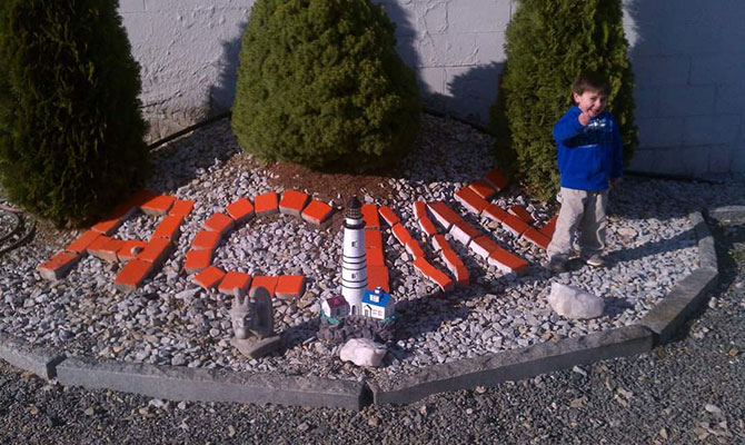 Child standing beside decorative stones spelling "ACME" in a landscaped area with shrubs and ornamental features, representing ACME Pressure Washing's commitment to community and service in Merrimack, NH.