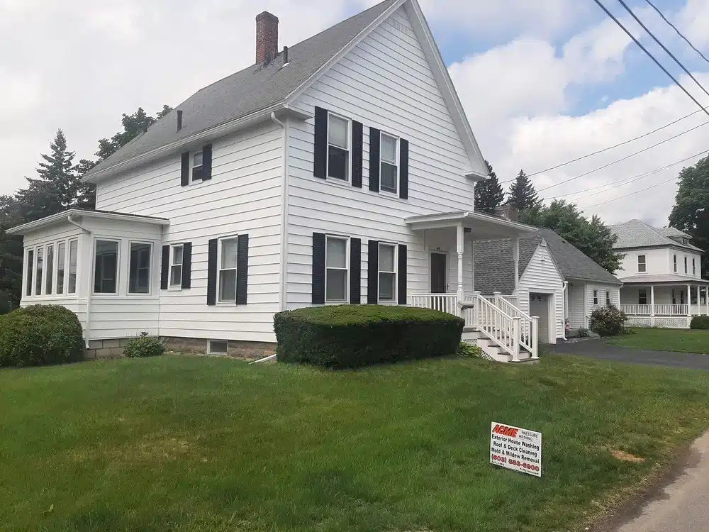 White two-story house with black shutters and a manicured lawn, featuring a sign for ACME Pressure Washing promoting exterior house cleaning services.