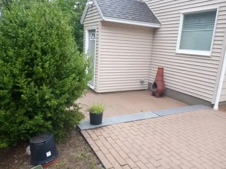 Brick patio with potted plants next to a house with white vinyl siding and a small gable structure