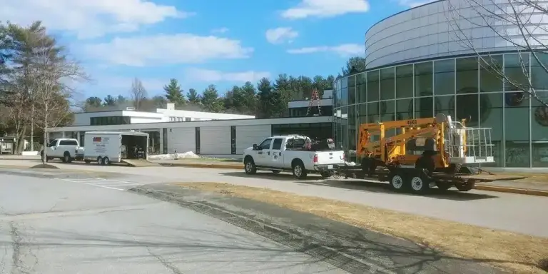 White pickup truck towing yellow pressure washing equipment on a commercial property with a modern building and storage tank