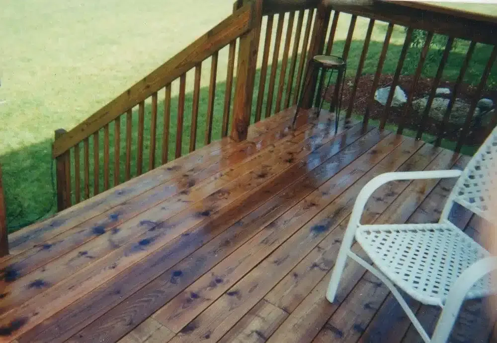 Wooden deck with visible discoloration and stains on the boards, brown wooden railing, and white plastic chair