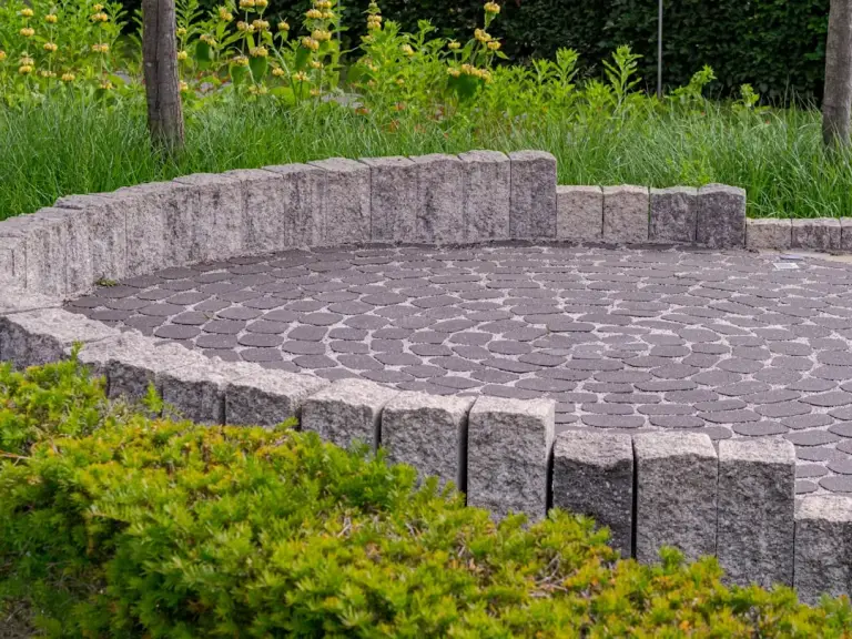 Curved patio with dark paver stones arranged in a spiral pattern, bordered by gray stone blocks and surrounded by green landscaping