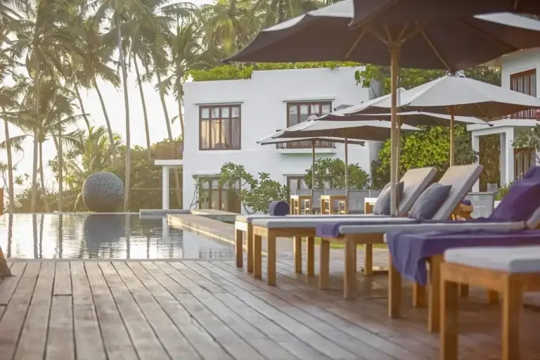 Poolside deck with wooden lounge chairs, umbrellas, and a white building with palm trees