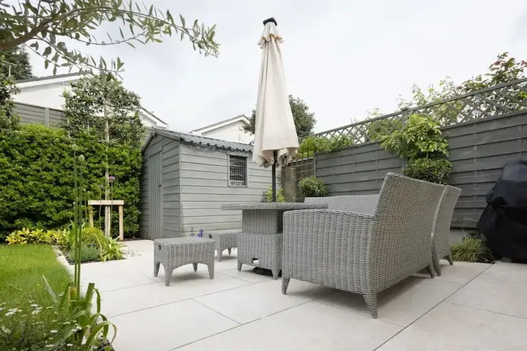 Clean patio with gray wicker furniture, umbrella, and surrounding garden beds enclosed by dark fencing